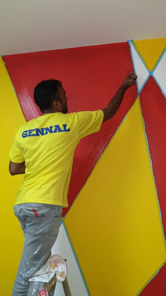 A man on a stepladder painting a red and yellow geometric accent wall using painter's tape.
