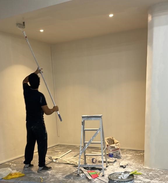 A painter applying white paint to a ceiling using an extension roller in a room undergoing renovation, with paint cans and a ladder on the floor.