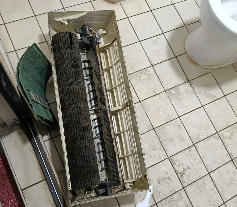 Disassembled dirty air conditioner housing and rotary blower wheel sitting on bathroom floor tiles next to a cleaning brush.